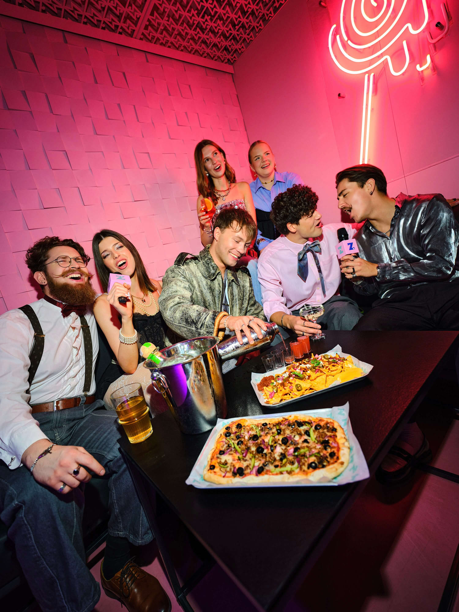 Grupa znajomych śpiewająca karaoke przy stole z pizzą i nachos w klimatycznym pomieszczeniu